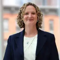 Abbie Eastman, a smiling woman with curly hair wearing a navy blazer over a white top
