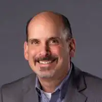 John Modesto, a smiling bald man with a moustache and goatee, wearing a grey jacket