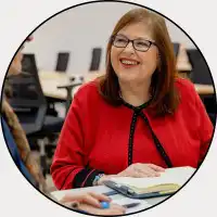 Anna McGovern, a smiling woman with glasses and a red top with black trim, seated in a room with a notebook