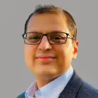 Mukul Agrawal, a smiling man with glasses wearing a light blue shirt and dark blazer against a grey wall