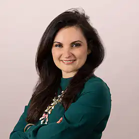 Alla Valente, a smiling woman with long dark hair wearing a green top and a floral necklace, arms crossed