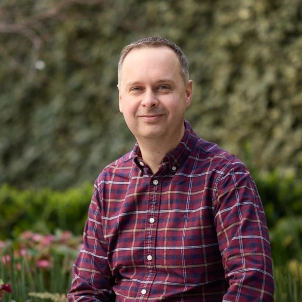 Dave Everett, a smiling man in a red and navy plaid shirt sitting in a garden with green hedges