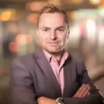 A man with arms crossed, in a grey jacket over a pink striped shirt, with blurred lights behind him