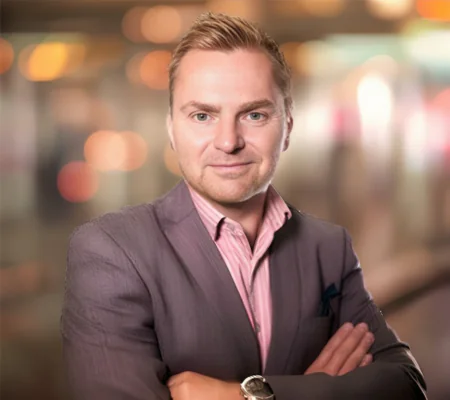 A man with arms crossed, in a grey jacket over a pink striped shirt, with blurred lights behind him